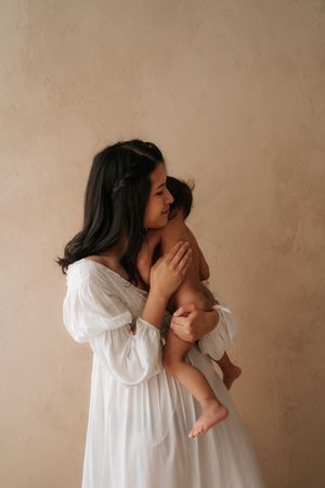 A mother in a white dress tenderly holds her newborn baby against a neutral beige wall.
