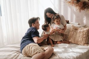 A happy Asian family with two toddlers sitting together on a cozy bed in a bright, sunlit room.