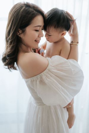 A young Asian mother in a white off-shoulder dress tenderly holds her baby, touching foreheads in a bright room.