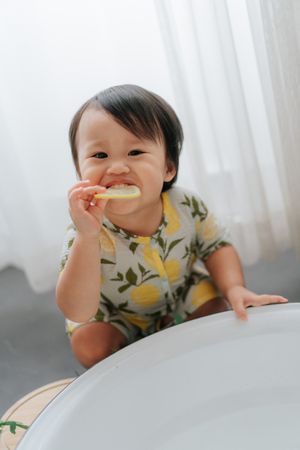 Cute Asian toddler in a lemon-print outfit smiling while chewing on a teether toy near a white surface.