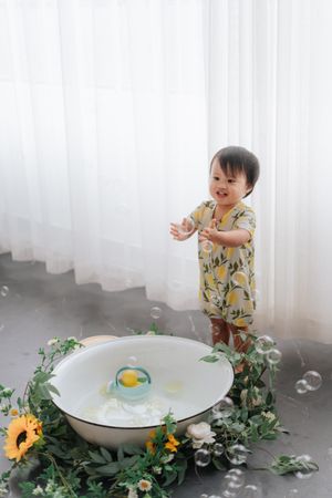 A smiling toddler in a yellow floral romper plays near a white basin with flowers and bubbles by sheer curtains.