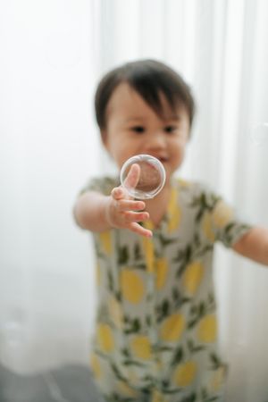 Toddler in yellow floral outfit holding a clear bubble toward camera with white curtain background.