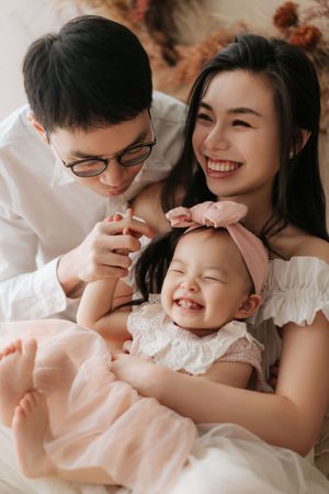 Happy Asian family with smiling parents and laughing baby girl in pink dress during a cozy photo session.
