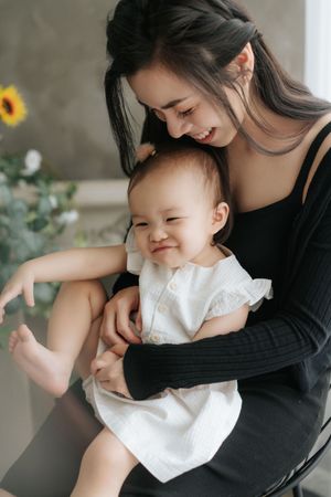 A smiling young Asian mother in black dress holds her happy baby girl in white dress near sunflowers.