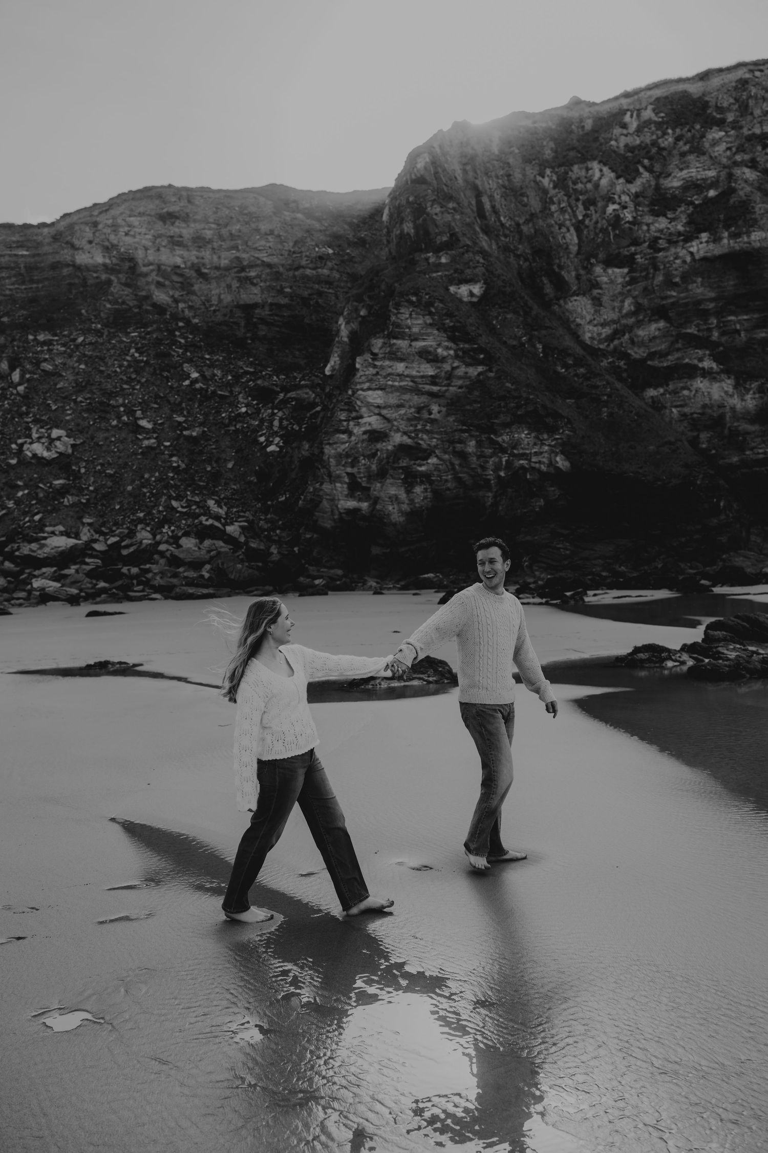 Black and white photo of a couple holding hands and walking barefoot at watergate bay with dramatic rocky cliffs behind them.