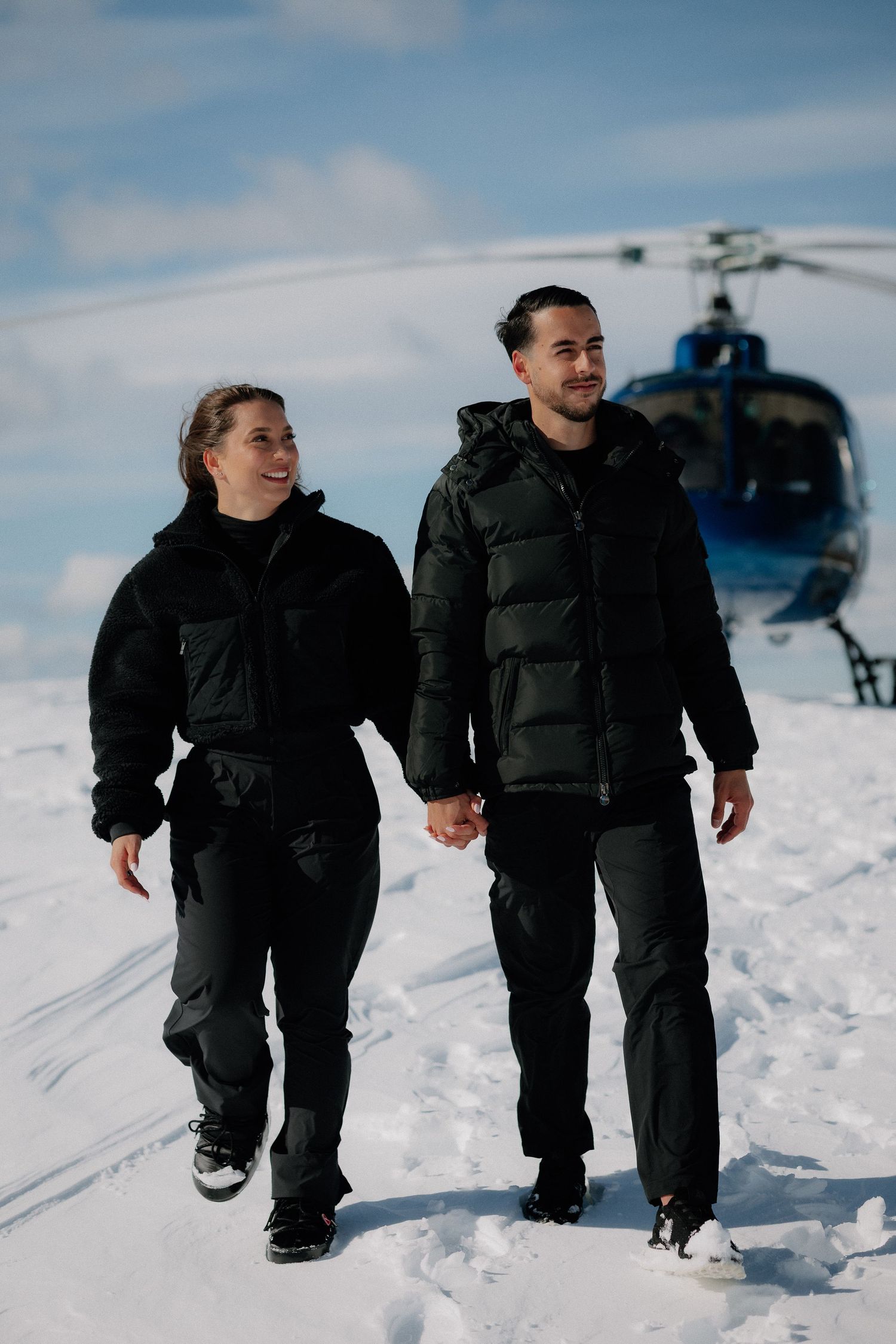 Couple standing on snowy mountain summit with panoramic winter landscape and cloudy sky in background.