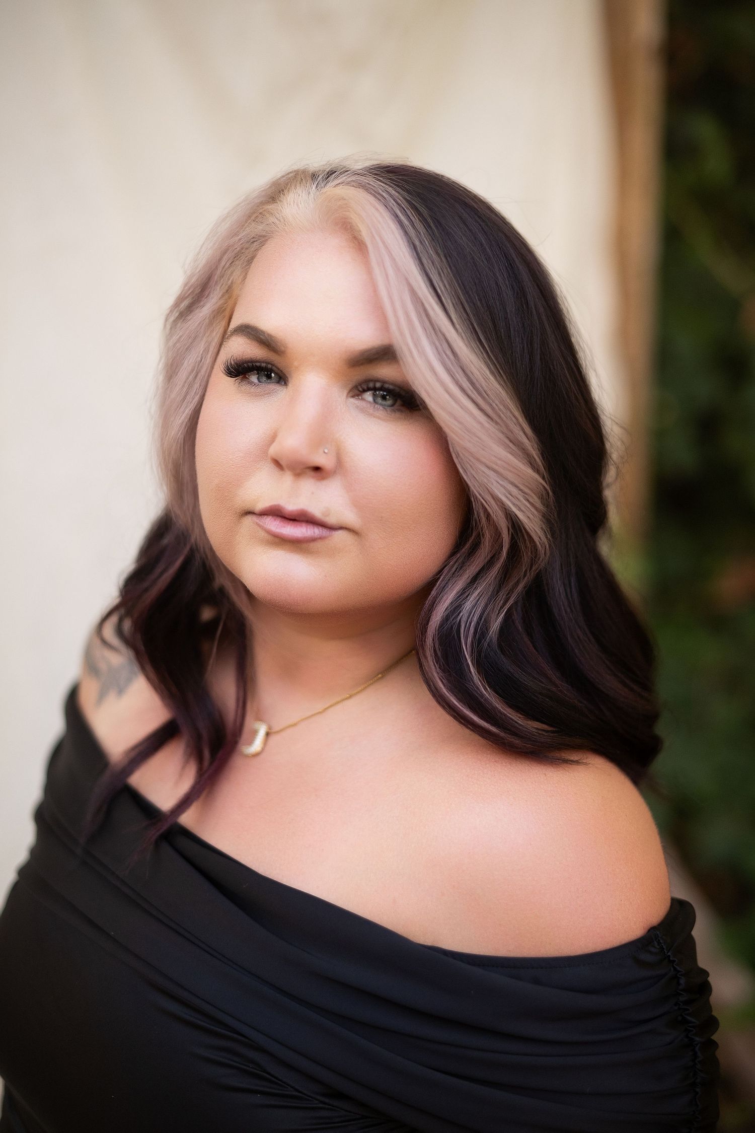 Woman with two-toned blonde and dark hair wearing black off-shoulder top and gold necklace poses outdoors.