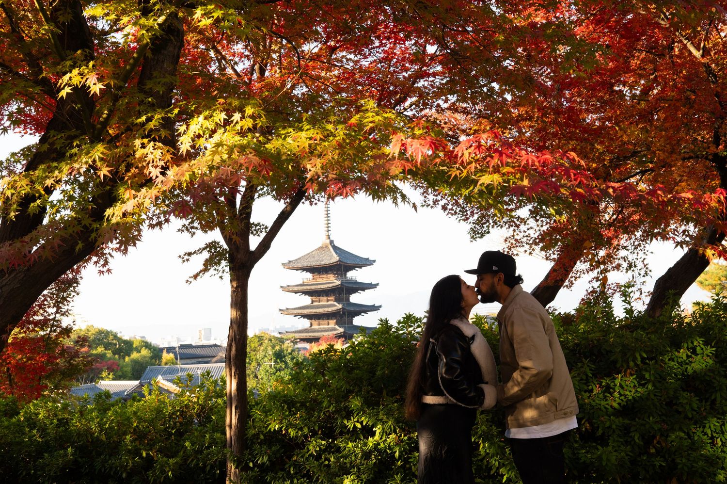 Couple kissing beneath vibrant red autumn maple leaves with a traditional Japanese pagoda rising in the background.