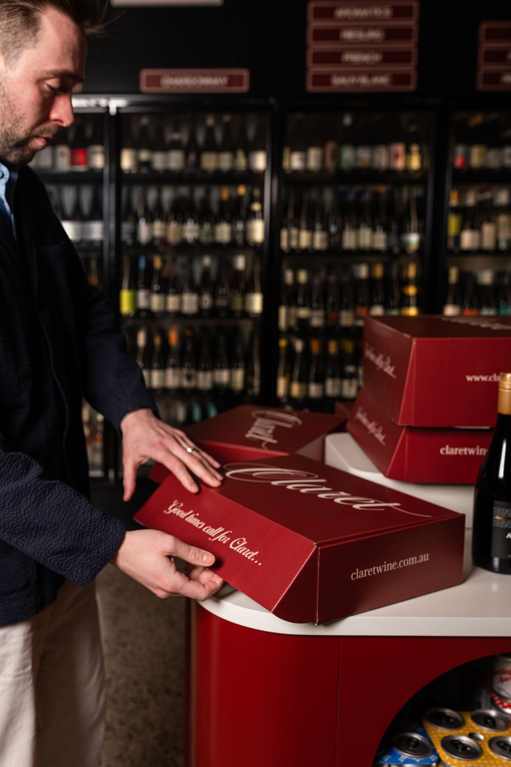 Man in a navy jacket closes a red Claret wine gift box on a red counter in a bottle shop with shelves of wine behind him.