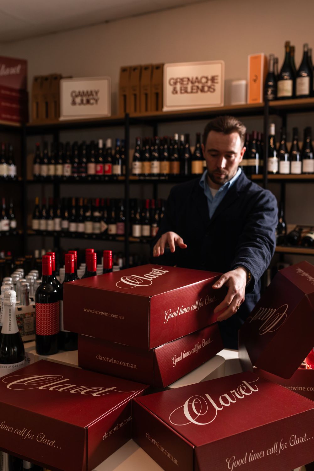 Man in a navy jacket stacks red Claret gift boxes in a wine shop with shelves of red wine bottles and Grenache & Blends signs.