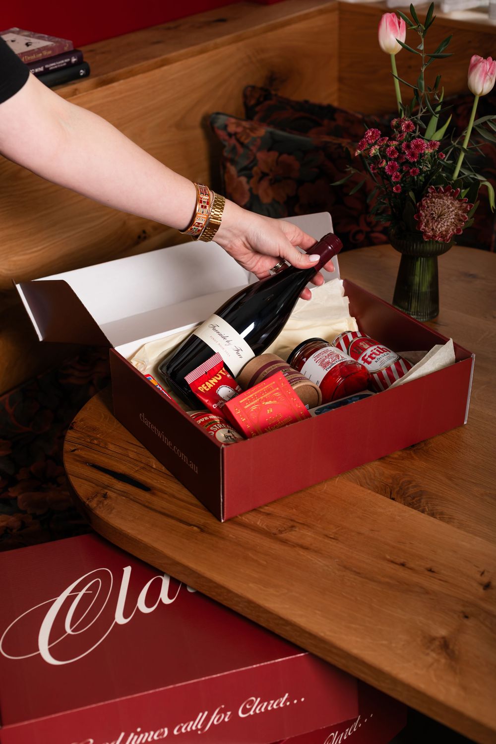 Hand placing a bottle of red wine into a burgundy Claret gift box with jars of preserves, set on a wooden table beside fresh pink tulips.