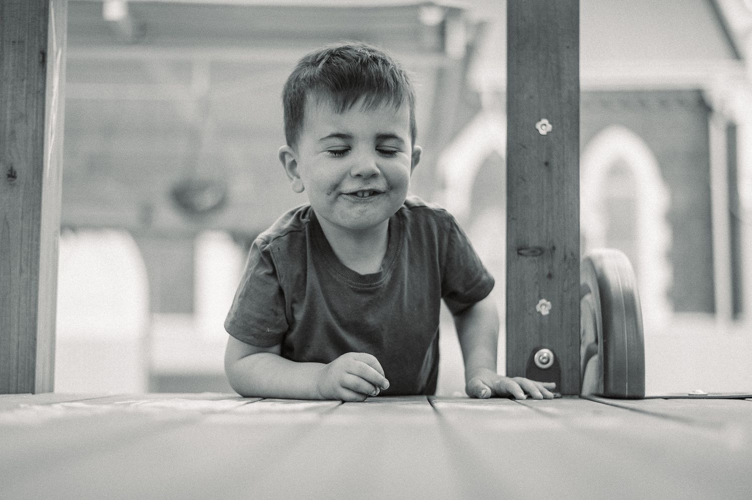 Toddler in a navy sweater plays with a metal pan and utensils at a wooden outdoor mud kitchen in a garden with a playground behind.