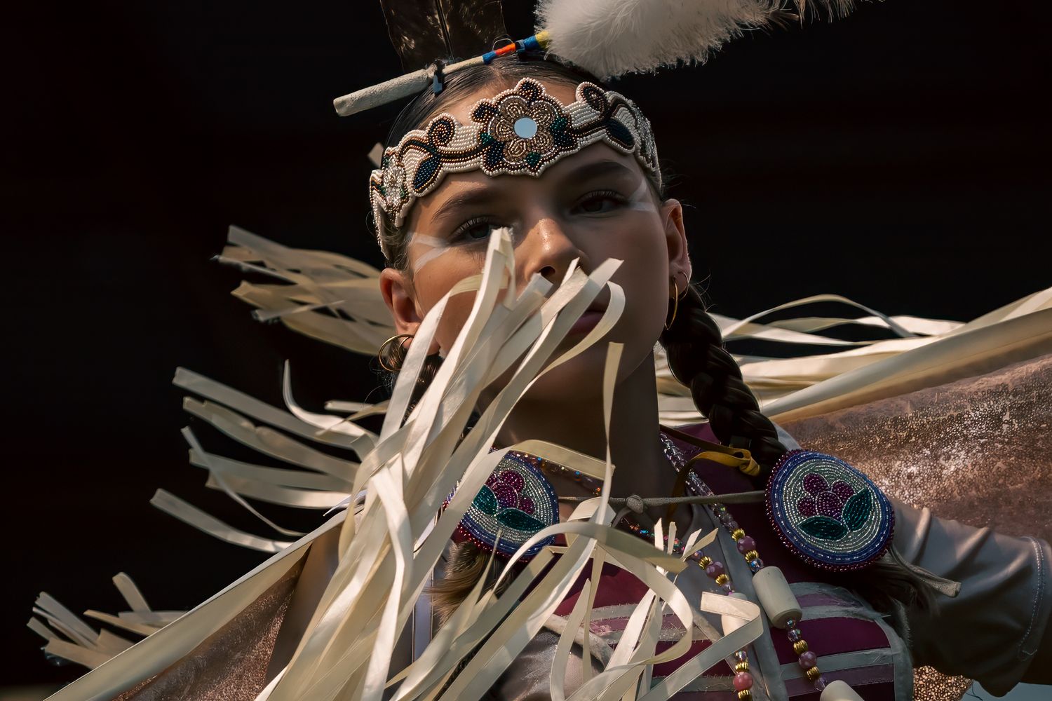Dancer in traditional regalia with a beaded headband, feather accent, long braid and flowing white fringe, moving against a dark background.