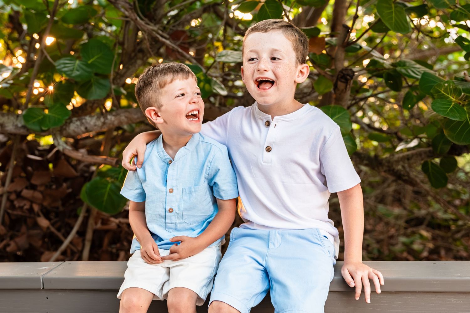 Two young boys laughing and hugging outdoors, wearing light blue outfits, seated together near tropical greenery.