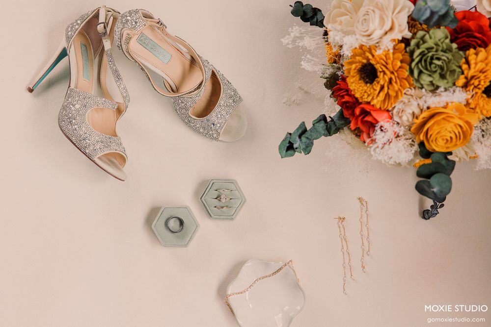 Bridal flat lay featuring glittery heels, floral bouquet, ring box, and gold hair pins on a blush background.