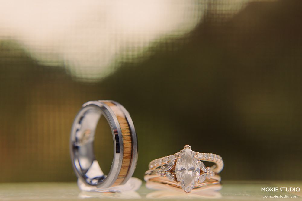 Close-up of a marquise diamond engagement ring in rose gold beside a tungsten wedding band with wood inlay on a reflective surface.