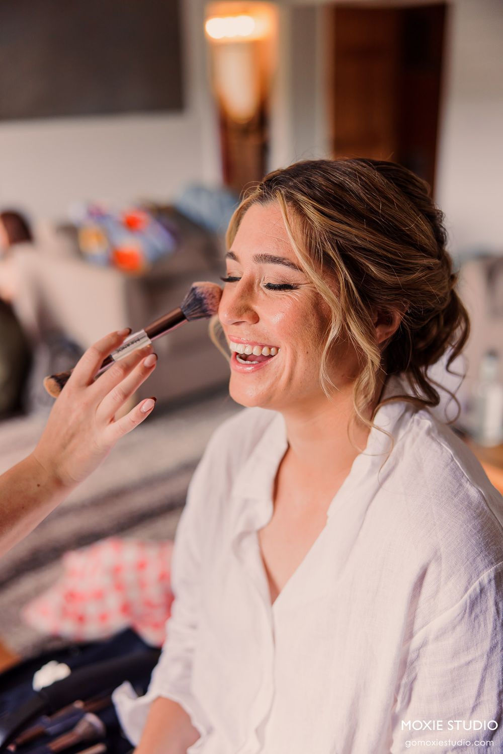 Bride smiling joyfully while getting makeup applied with a brush during wedding day preparations.