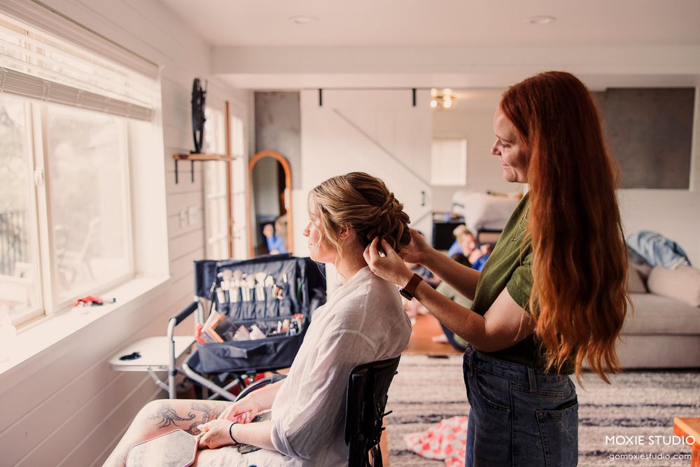 A redheaded stylist does hair for a seated woman near a bright window in a modern home studio.