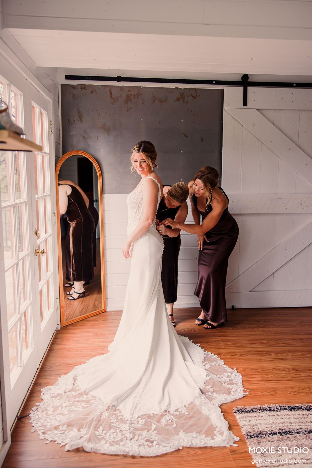 Bride in elegant lace gown being helped by bridesmaids in rustic barn venue with hardwood floors.