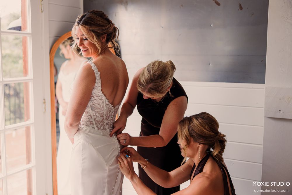Bride being helped into her wedding dress by two women in a bright room.