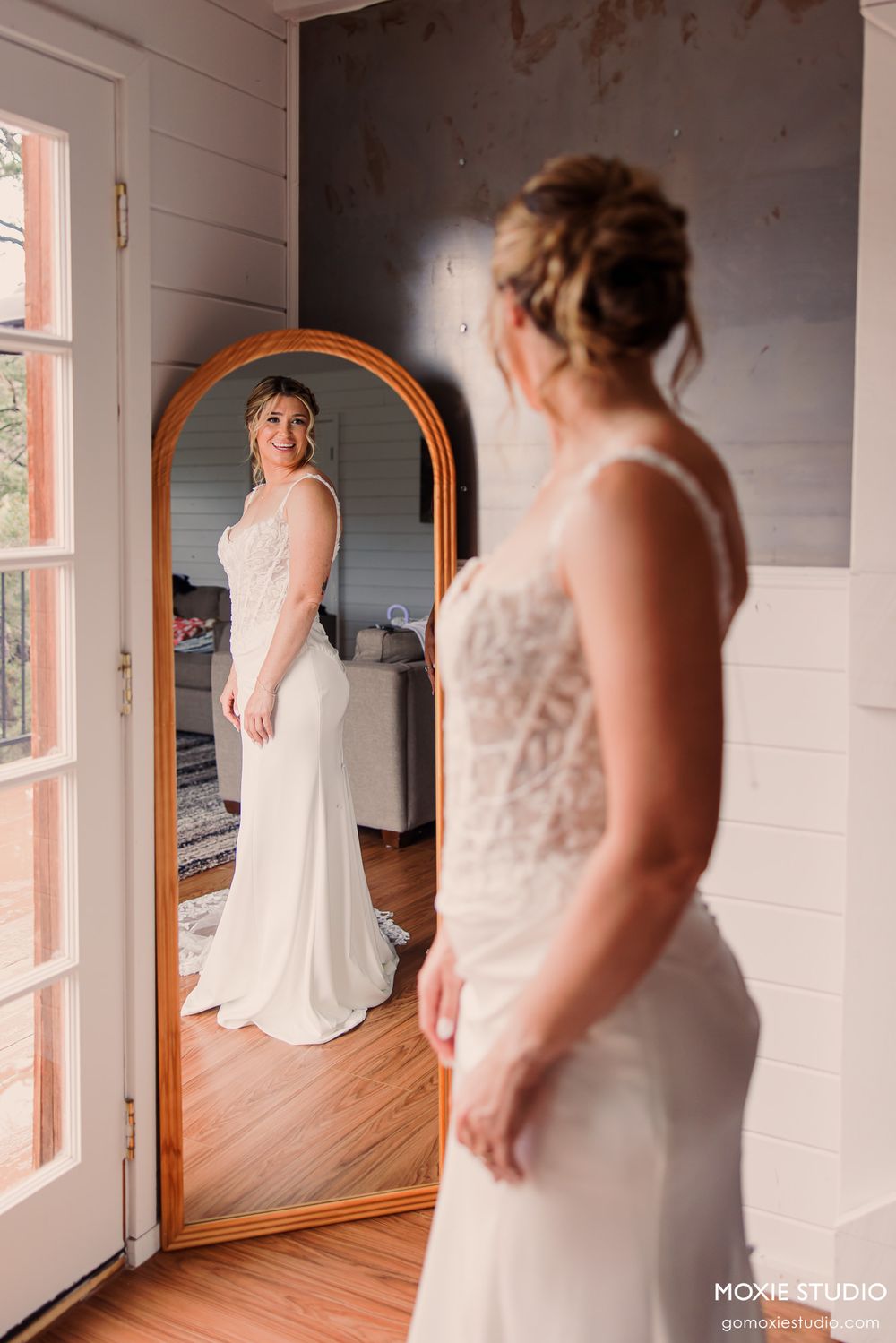 Bride in white lace wedding gown admires her reflection in an arched floor mirror during bridal prep.