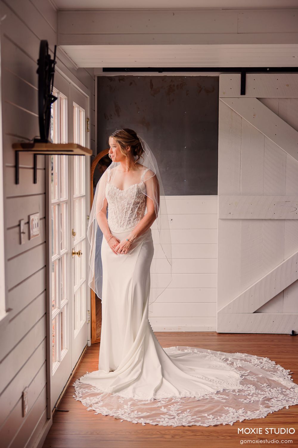 Elegant bride in lace-trimmed veil and fitted gown standing by window in rustic barn venue.