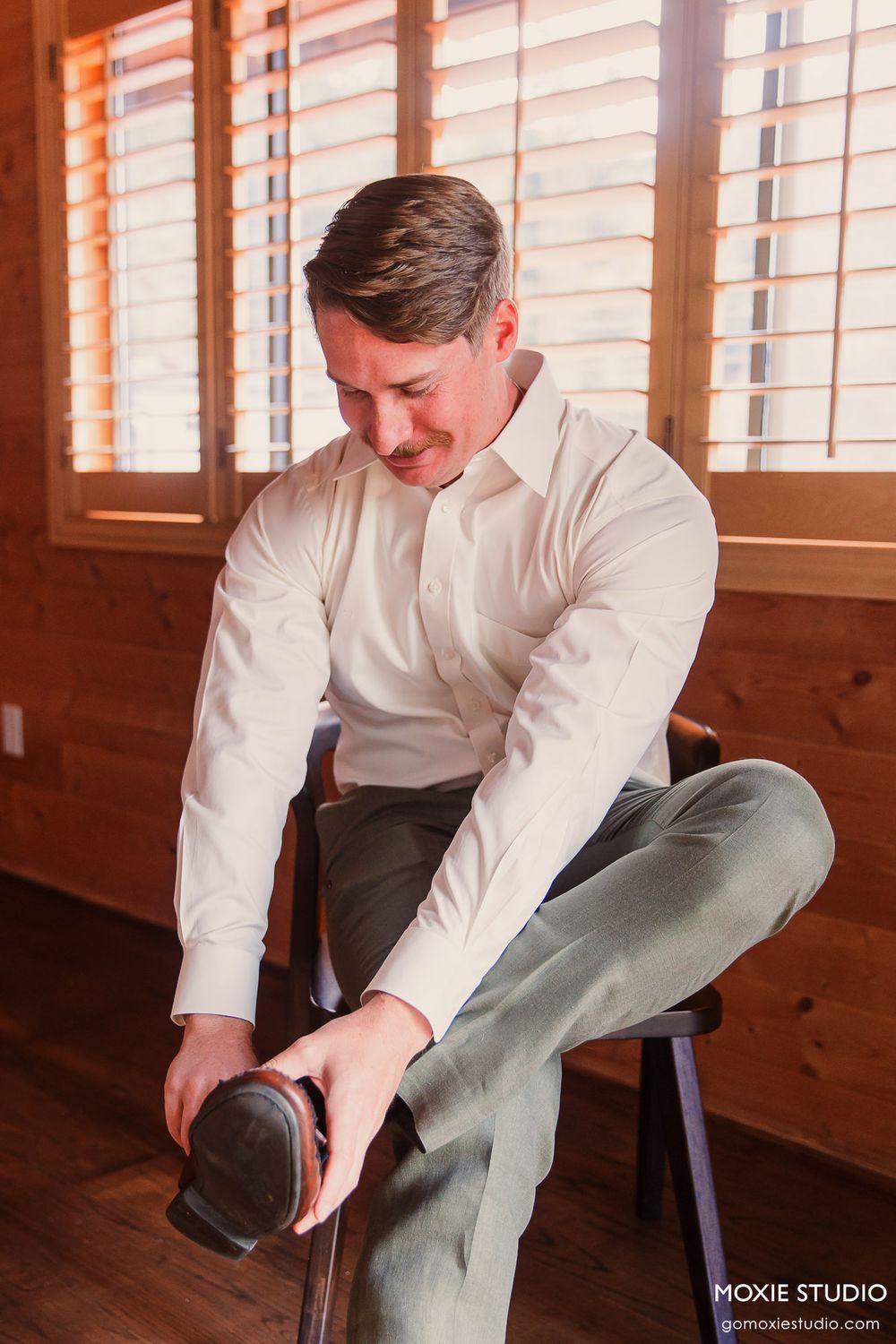 Groom in white dress shirt and gray pants putting on dress shoes while seated on a wooden chair near shuttered windows.