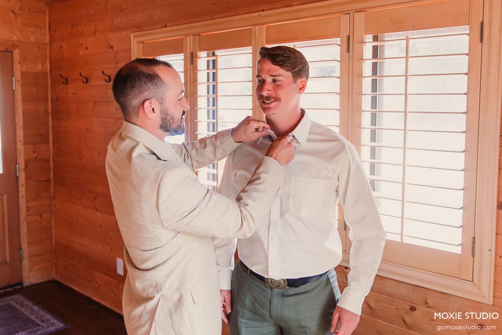 Groomsman helping groom adjust his tie while getting ready in a rustic wooden cabin before the wedding.
