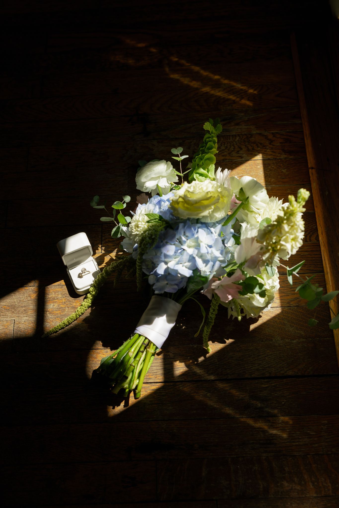 Bridal bouquet of white and blue hydrangeas with greenery beside a ring box on rustic wooden floor in natural light.