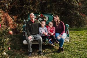 Family of four sitting on the tailgate of a vintage green pickup truck decorated with holiday greenery in an autumn outdoor setting.