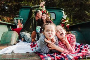 Two smiling girls in cozy sweaters lie on a plaid blanket in a vintage green truck bed while their parents kiss behind them at Christmas.