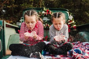 Two girls with braided hair sit in a vintage truck bed, blowing fake snow from their hands during a festive Christmas photo shoot.