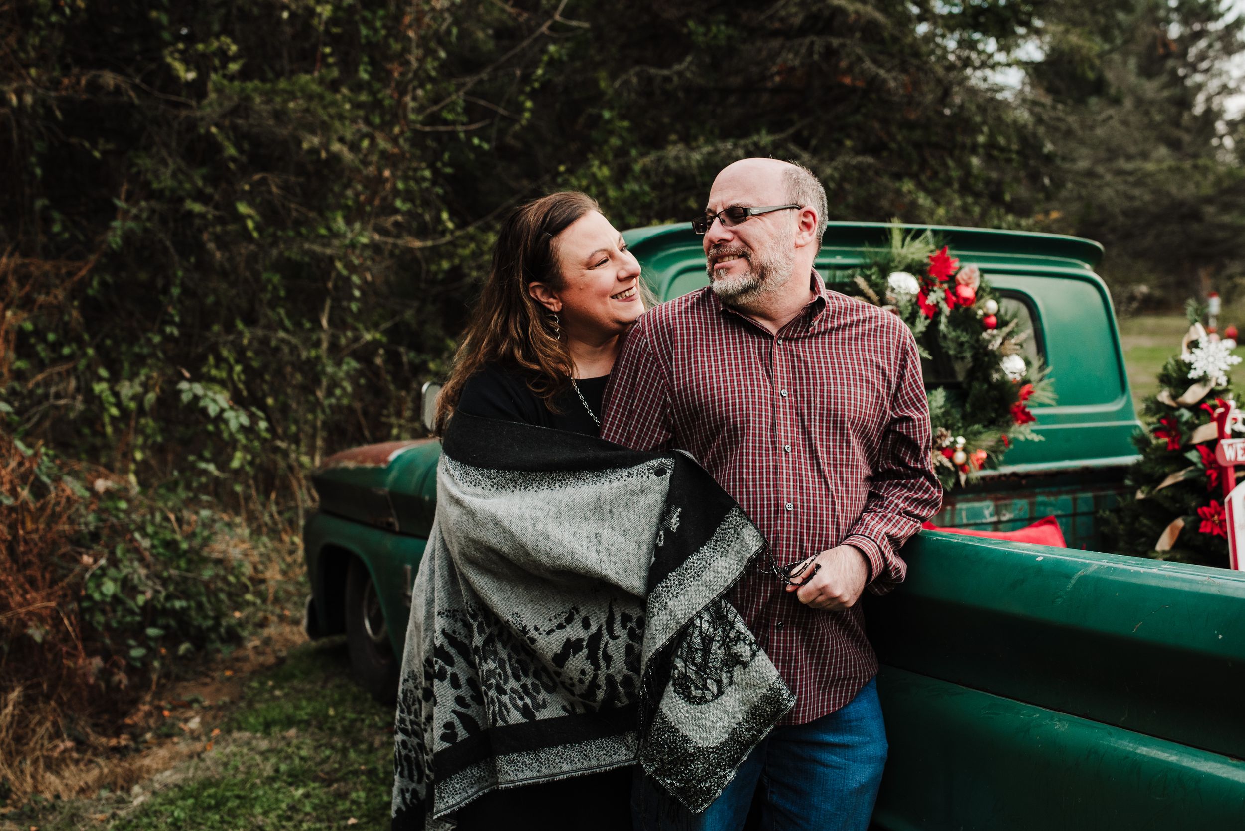 Smiling couple embracing beside a vintage green truck decorated with a red-flowered Christmas wreath during a holiday family photo session.