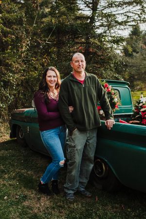 Smiling couple poses beside a vintage green pickup truck decorated with Christmas flowers and greenery in a sunlit evergreen forest.