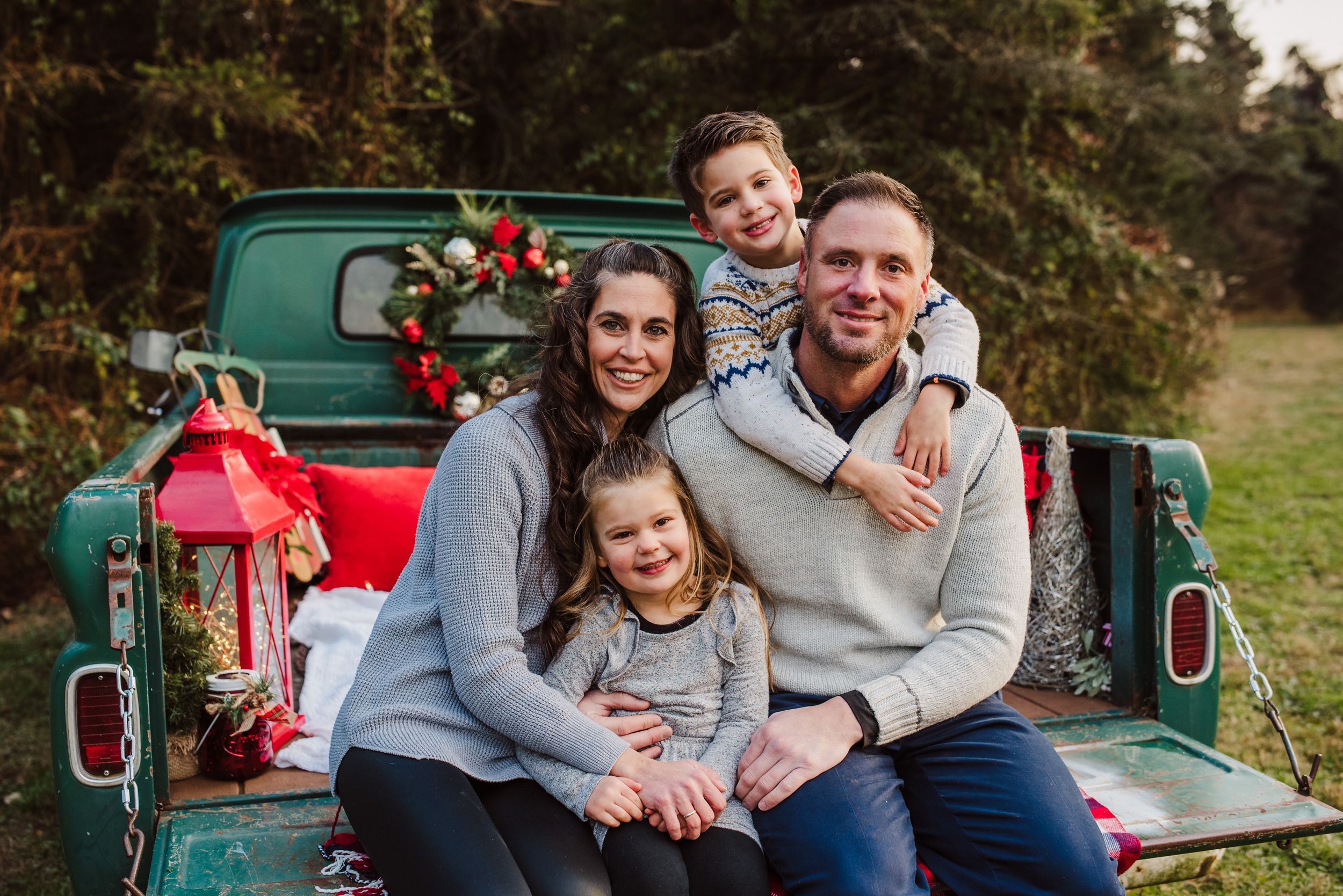 Smiling family of four sitting on the tailgate of a vintage green pickup truck decorated with a red lantern and holiday greenery.