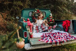 Woman and child sit on the tailgate of a vintage green pickup truck decorated with Christmas wreaths, plaid blankets and red lanterns.