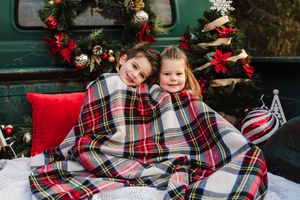 Two smiling children wrapped in a red plaid blanket sit in a vintage truck bed decorated with Christmas wreaths, red bows and ornaments.