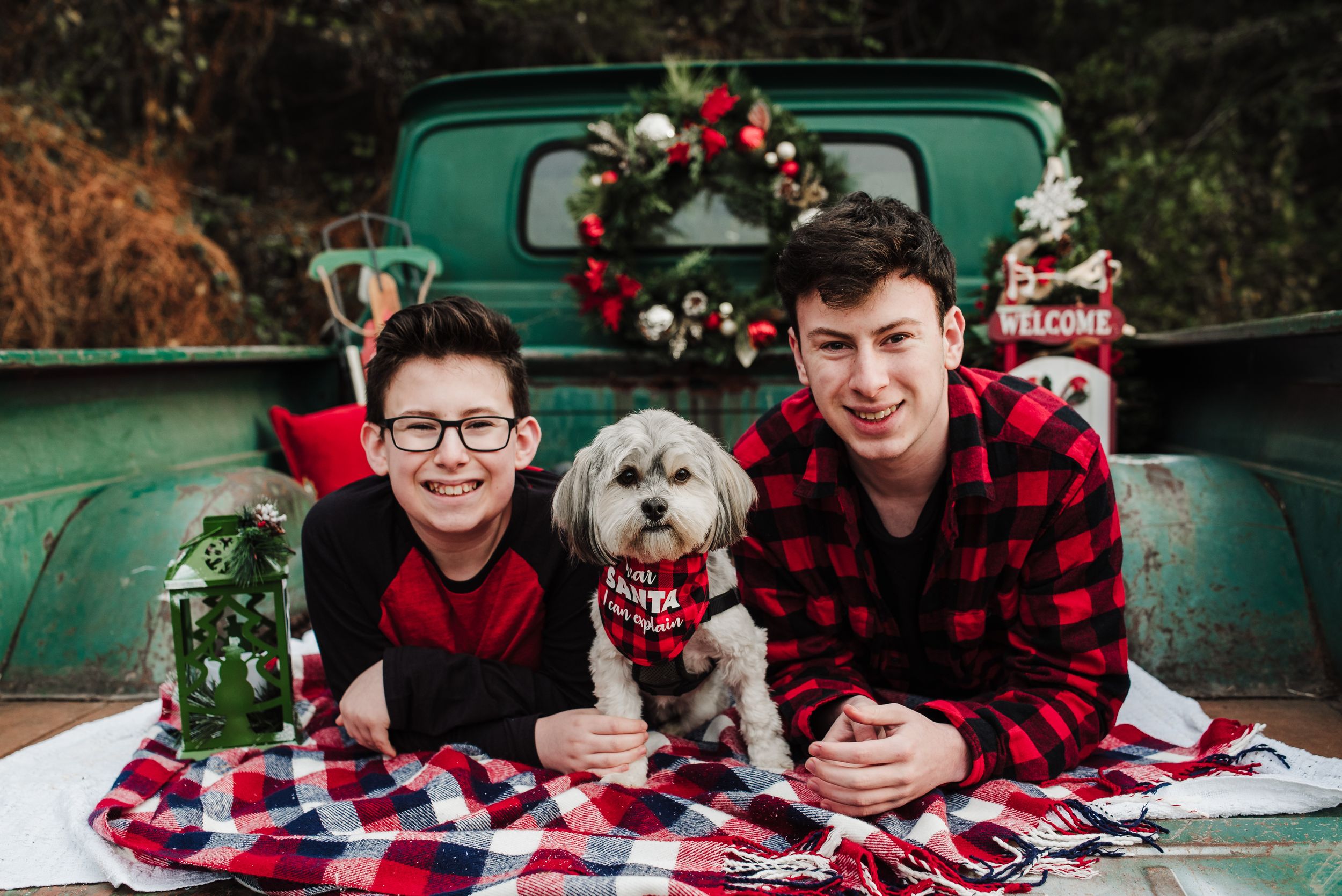 Two smiling boys in red plaid and a fluffy white dog pose on a blanket in front of a vintage green truck with a Christmas wreath.