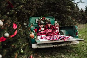 Family wrapped in red plaid blankets sits on the tailgate of a vintage green pickup truck decorated with a Christmas wreath.
