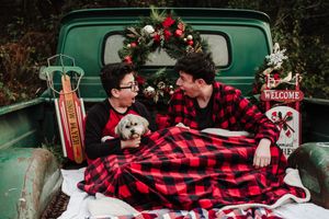 Two young men sit under a red buffalo plaid blanket with a small dog in a vintage green pickup truck bed decorated with a Christmas wreath.