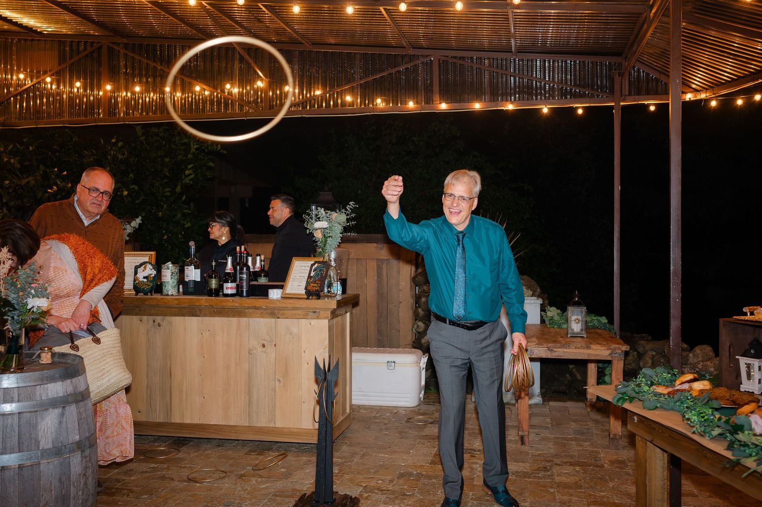 Man in teal shirt raises hoop at outdoor evening party with string lights and bar in background.