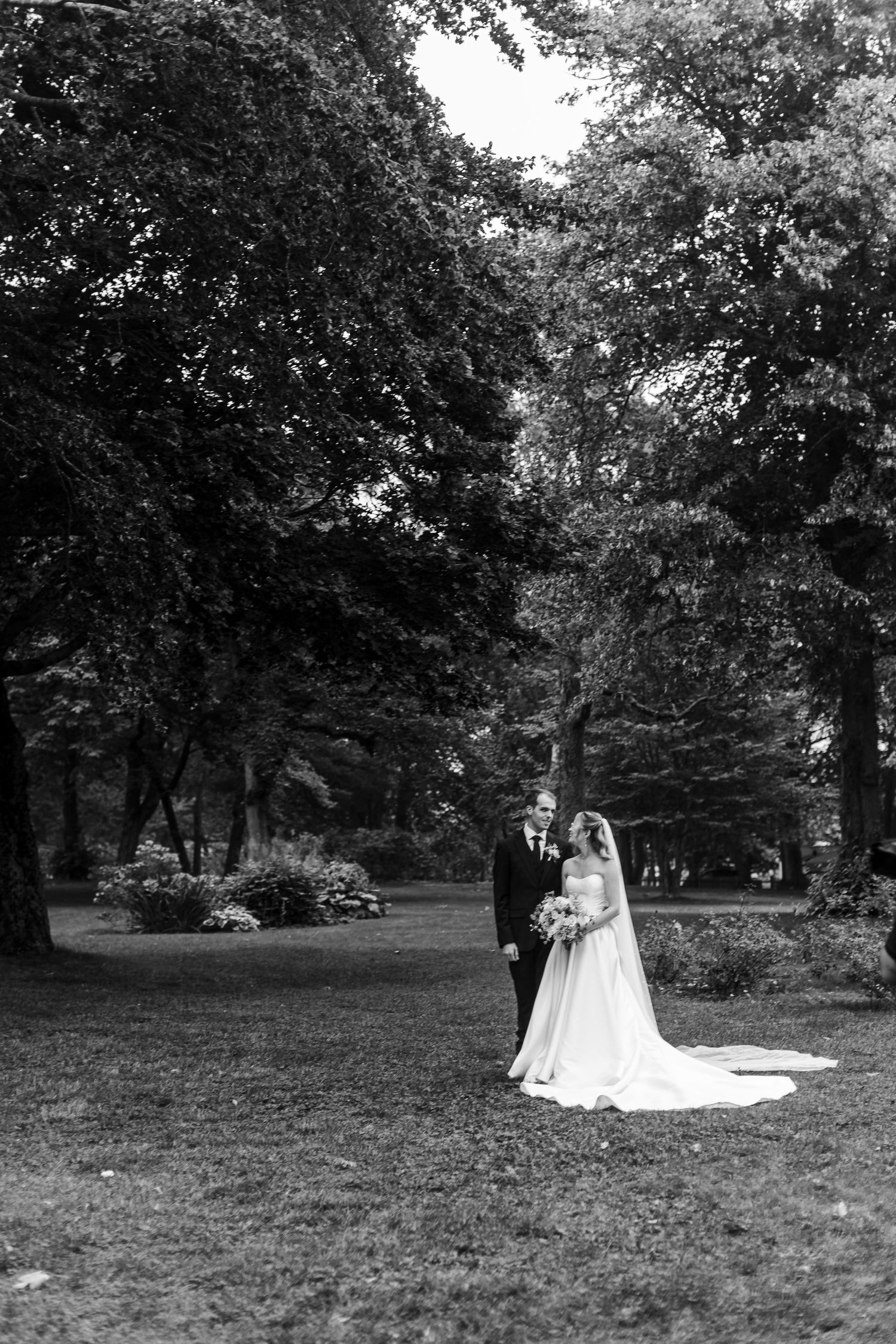 Black and white photo of a bride and groom standing together in a lush green park on their wedding day.