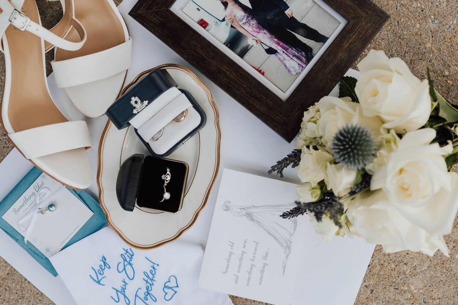 Wedding flat lay with white heels, rings, bouquet of white roses, framed photo, and blue calligraphy stationery.