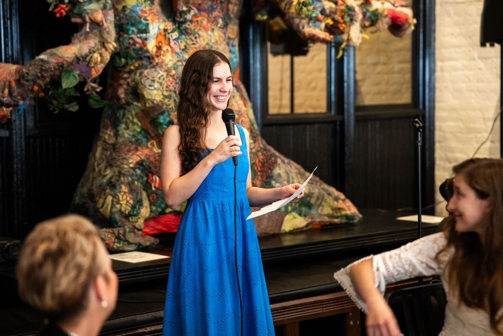 Woman in blue dress speaks into microphone at colorful indoor event.