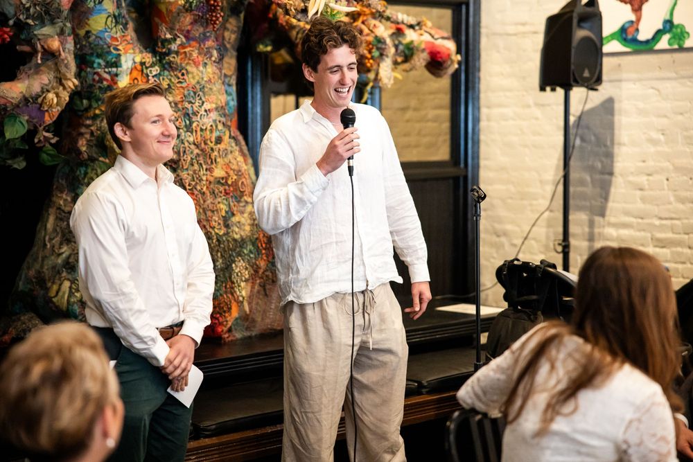 Two men in white shirts share microphone at vibrant indoor gathering.