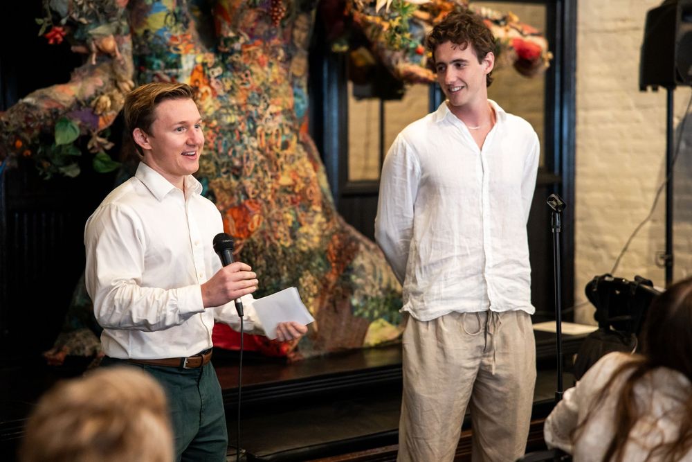 Two men in white shirts speaking at an indoor event with colorful floral decor in the background.