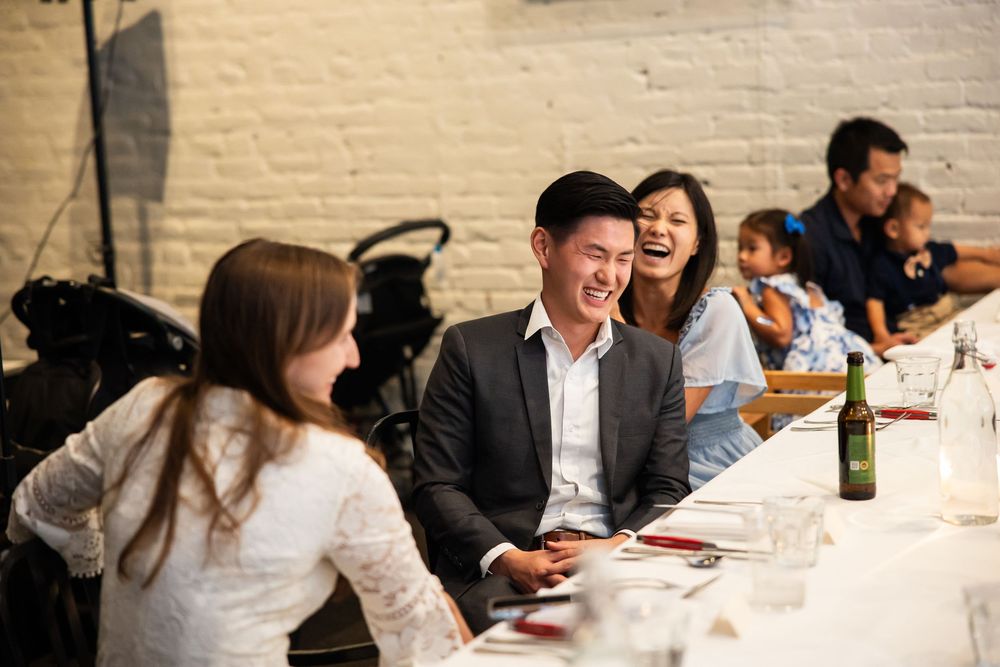 People laughing and socializing at a long dining table during an indoor dinner event in a white brick restaurant.