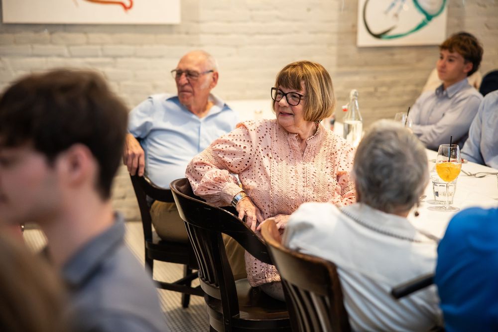 Family gathering around a restaurant dining table, smiling woman in pink blouse with glasses enjoying conversation with elderly guests.