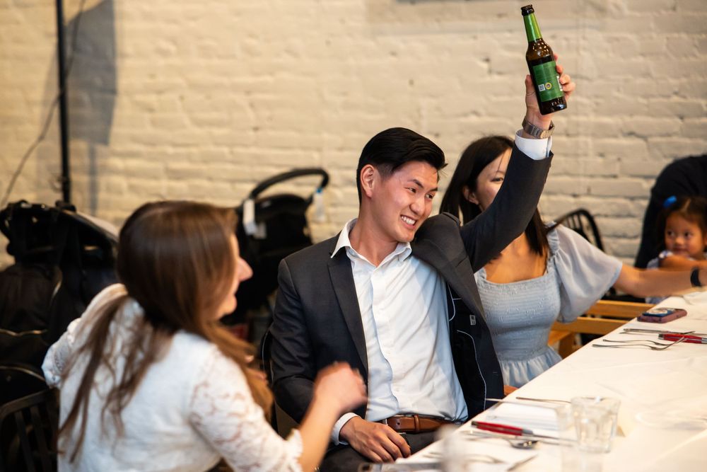 A smiling man in a suit raises a green beer bottle at a restaurant dinner table with guests seated around him.
