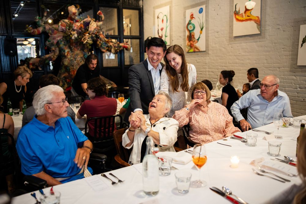 Guests enjoying a celebratory dinner at an elegant restaurant, with a couple greeting elderly diners at a white tablecloth table.